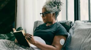 Woman reading a book in bed with a CGM device on her arm.